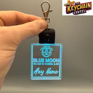 May include: A light-up keychain with a square, transparent design. The keychain features the text "BLUE MOON" and "Any Name" in a blue glow. The top of the keychain has a black base and a silver clasp. The logo "THE KEYCHAIN CARTEL" is in the upper right corner.