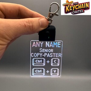 May include: A clear acrylic keychain with the text "ANY NAME SENIOR COPY-PASTER" and keyboard shortcut symbols. The keychain is held by a silver clip and a black attachment. The Keychain Cartel logo is in the upper right corner.