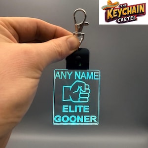 Könnte beinhalten: Ein leuchtender, quadratischer Schlüsselanhänger mit dem Text "ANY NAME" über einer Faustgrafik und den Worten "ELITE GOONER" darunter. Der Schlüsselanhänger wird von einem silbernen Clip und einer schwarzen Basis gehalten. Das Keychain Cartel-Logo befindet sich in der oberen rechten Ecke.