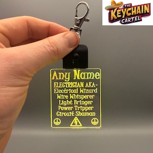 May include: A yellow LED keychain with the text "Any Name ELECTRICIAN AKA- Electrical Wizard Wire Whisperer Light Bringer Power Tripper Circuit Shaman" and a warning sign. The keychain has a silver metal ring and a black button.