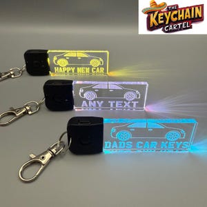 May include: Three illuminated car keyrings with different text: "HAPPY NEW CAR", "ANY TEXT", and "DADS CAR KEYS". Each keyring has a black remote and a silver clasp. The keyrings are rectangular and feature a car silhouette.