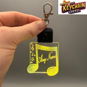 May include: A clear, illuminated keychain with a musical note design. The note is outlined in yellow and features the text "Any Name" in a script font. The keychain includes a silver clasp and a black light component. The logo "The Keychain Cartel" is in the upper right corner.