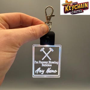 Puede incluir: Un llavero cuadrado e iluminado con un cierre plateado. El llavero presenta el texto "I'm Forever Blowing Bubbles" y "Any Name" debajo de un gráfico de martillos cruzados. El logotipo de The Keychain Cartel está en la esquina superior derecha.