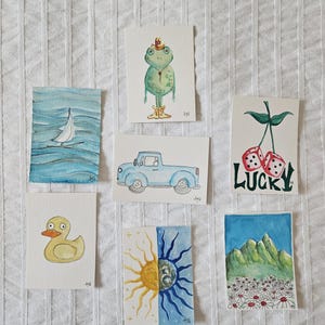 May include: A collection of watercolor art prints featuring various whimsical illustrations. The prints include a sailboat on blue water, a frog wearing a hat, a blue pickup truck, a yellow duck, a sun and moon design, dice with the word "LUCKY", and a mountain scene.