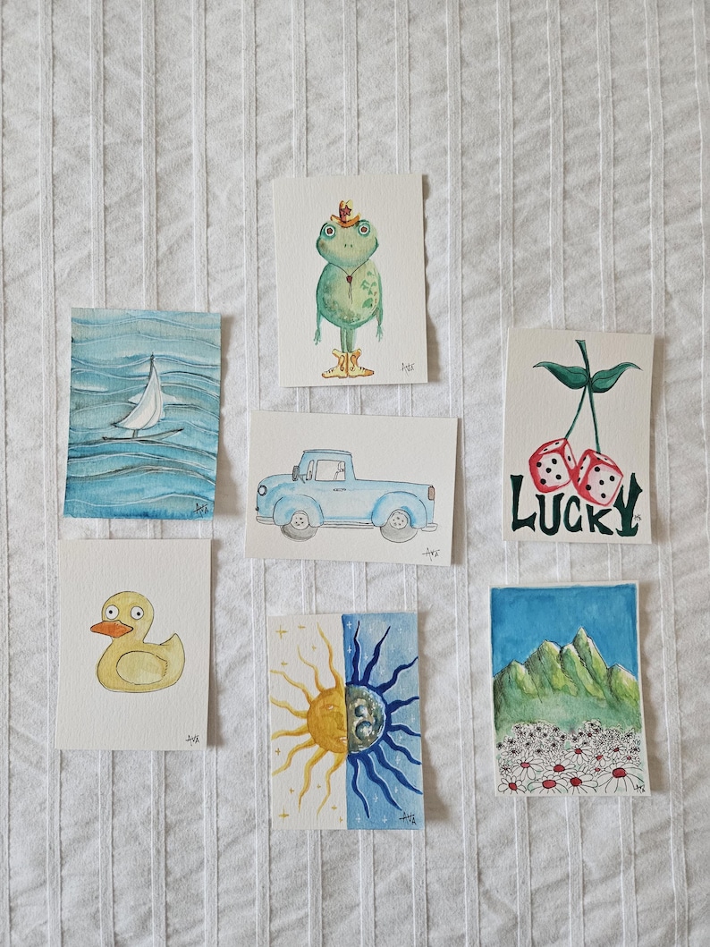 May include: A collection of watercolor art prints featuring various whimsical illustrations. The prints include a sailboat on blue water, a frog wearing a hat, a blue pickup truck, a yellow duck, a sun and moon design, dice with the word "LUCKY", and a mountain scene.