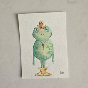 May include: Watercolor painting of a green frog wearing a cowboy hat and boots. The frog has large, round eyes and a small red bolo tie. The artwork is on a rectangular white paper.