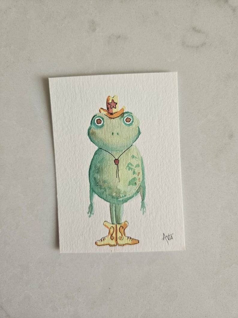 May include: Watercolor painting of a green frog wearing a cowboy hat and boots. The frog has large, round eyes and a small red bolo tie. The artwork is on a rectangular white paper.