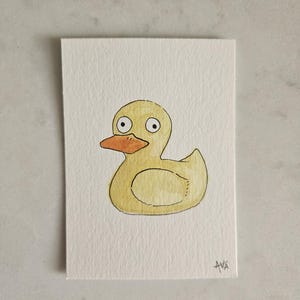 May include: A watercolor painting of a yellow rubber duck with an orange beak and large, round eyes. The duck is centered on a white rectangular card. The artist's signature, "AVA", is in the bottom right corner.
