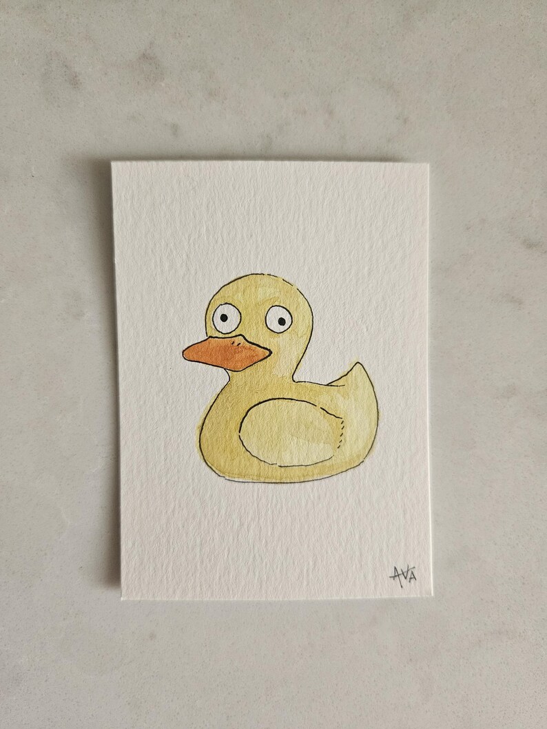 May include: A watercolor painting of a yellow rubber duck with an orange beak and large, round eyes. The duck is centered on a white rectangular card. The artist's signature, "AVA", is in the bottom right corner.
