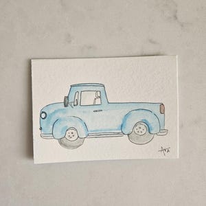 May include: Watercolor painting of a light blue vintage pickup truck. The truck has a rounded cab, a cargo bed, and gray wheels. The artwork is signed "Ava" in the lower right corner. The painting is on a white rectangular paper.