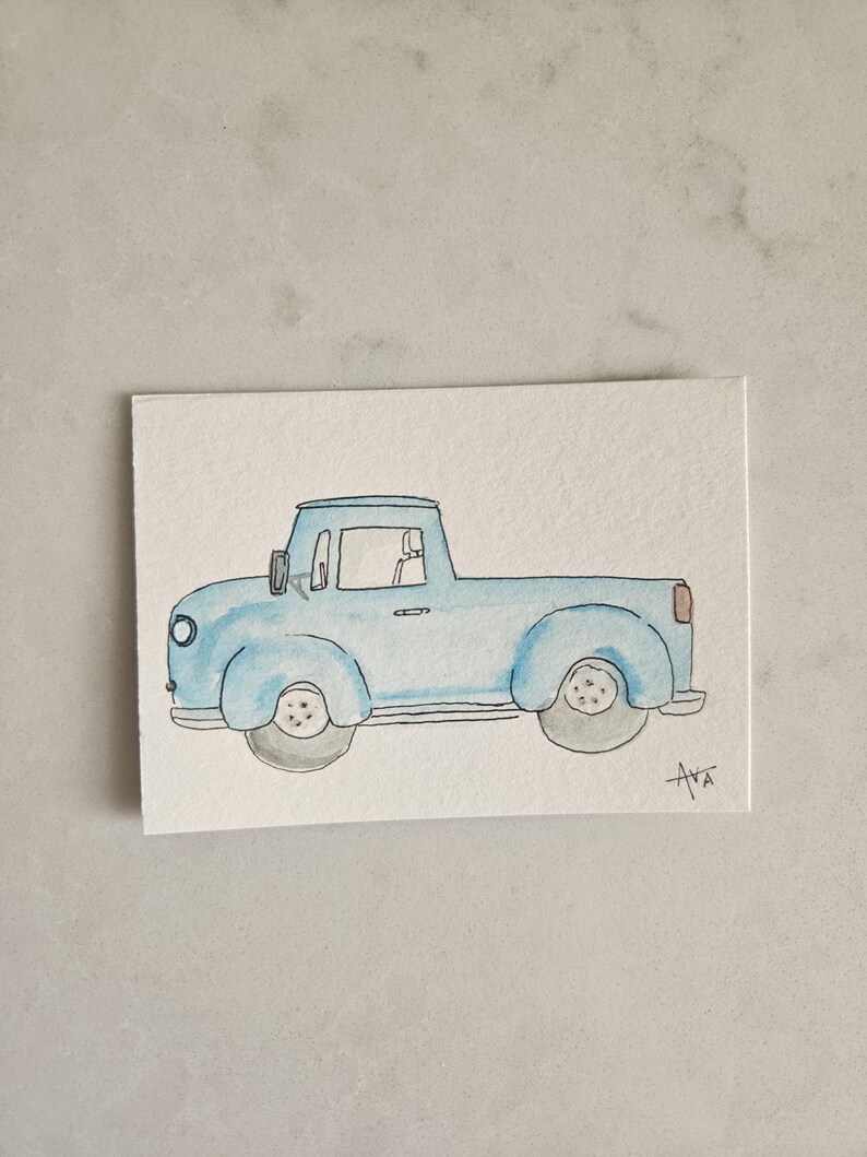 May include: Watercolor painting of a light blue vintage pickup truck. The truck has a rounded cab, a cargo bed, and gray wheels. The artwork is signed "Ava" in the lower right corner. The painting is on a white rectangular paper.