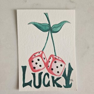 May include: Watercolor painting of two red dice with black dots, resembling cherries, connected by a green stem and leaves. The word "LUCKY" is written in green below the dice, creating a playful and whimsical design.