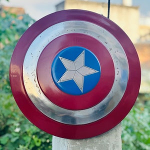K&ouml;nnte beinhalten: Ein rot-wei&szlig;-blauer Captain America Schild mit einem silbernen Stern in der Mitte. Der Schild h&auml;ngt an einer Schnur und befindet sich vor einem gr&uuml;nen Busch.
