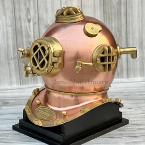 Peut inclure: Casque de plongée en cuivre et laiton, avec un corps rond et riveté, des hublots et des raccords. Monté sur une base en bois noir. Le texte "Iron Clad Armory" est visible.