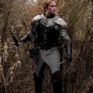 May include: A woman in a forest setting, wearing chainmail armor and a leather belt with a sword strapped to her leg. She is holding a dagger in her right hand and has a serious expression on her face.