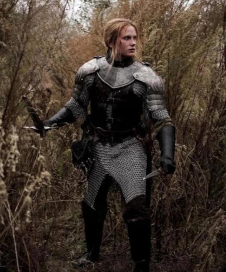 May include: A woman in a forest setting, wearing chainmail armor and a leather belt with a sword strapped to her leg. She is holding a dagger in her right hand and has a serious expression on her face.