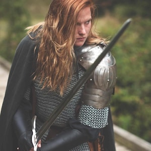 May include: A woman in chainmail armor and a black cloak holds a silver sword in front of her. She has long, red hair and is looking intently at something off-camera.
