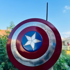 K&ouml;nnte beinhalten: Ein rot-wei&szlig;-blauer Captain America Schild mit silbernem Rand. Der Schild ist auf einem Betonpfosten aufgestellt.
