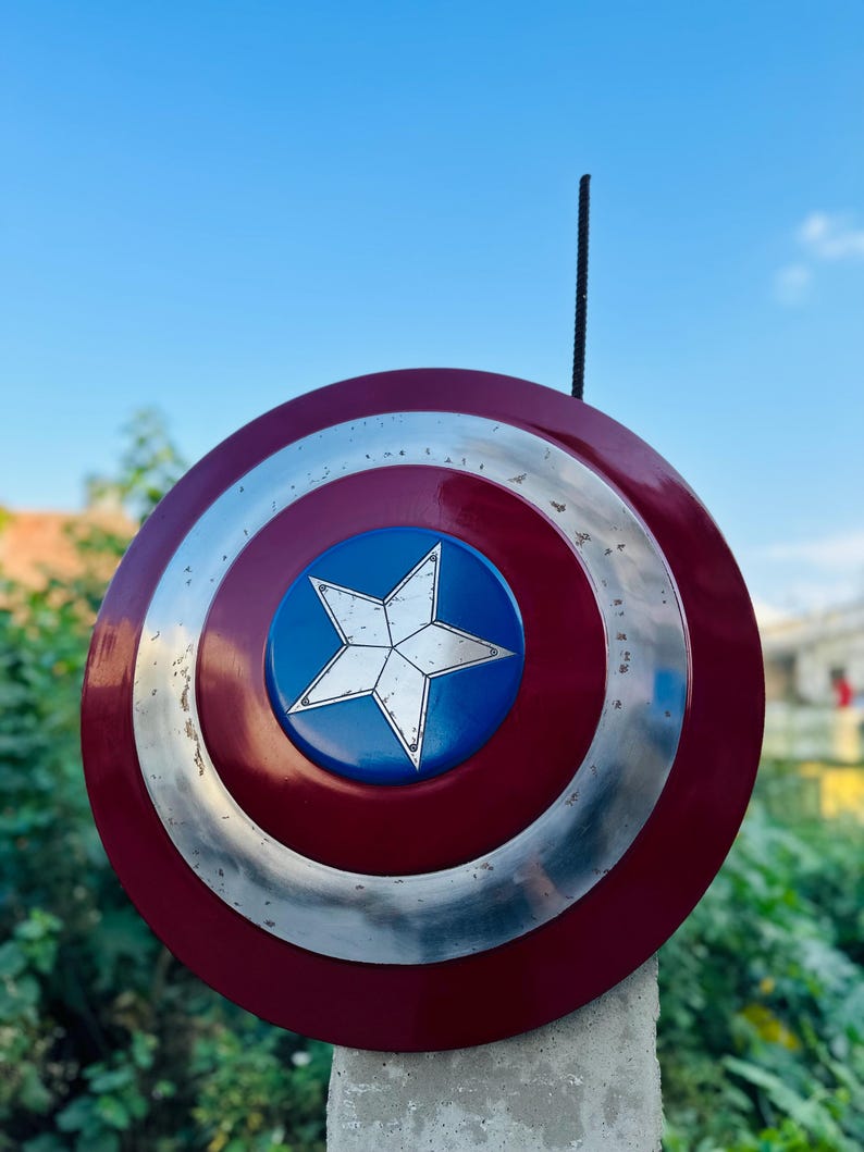 K&ouml;nnte beinhalten: Ein rot-wei&szlig;-blauer Captain America Schild mit silbernem Rand. Der Schild ist auf einem Betonpfosten aufgestellt.