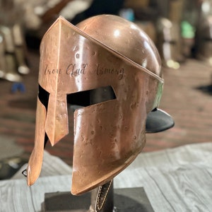May include: A copper-colored Spartan helmet with a black leather chin strap. The helmet is engraved with the words "Iron Clad Armory".