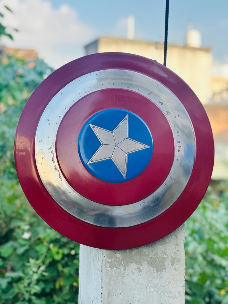 K&ouml;nnte beinhalten: Ein rot-wei&szlig;-blauer Captain America Schild mit einem silbernen Stern in der Mitte. Der Schild h&auml;ngt an einer Schnur und befindet sich vor einem gr&uuml;nen Busch.