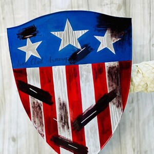 May include: A red, white, and blue shield with a distressed paint finish. The shield has a blue top with three white stars and a red and white striped bottom. The shield is painted with black brushstrokes. The text "Iron Clad Armory" is written in black on the shield.