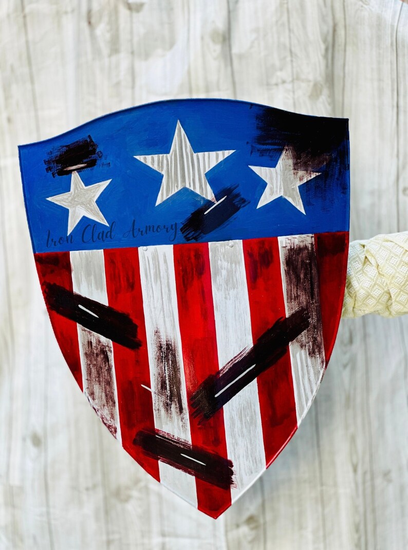 May include: A red, white, and blue shield with a distressed paint finish. The shield has a blue top with three white stars and a red and white striped bottom. The shield is painted with black brushstrokes. The text "Iron Clad Armory" is written in black on the shield.