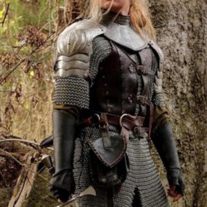 May include: A woman in a forest setting wearing chainmail armor, a leather jerkin, and a sword. She is wearing a brown leather belt with a pouch attached to it.