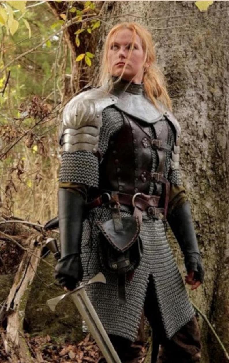 May include: A woman in a forest setting wearing chainmail armor, a leather jerkin, and a sword. She is wearing a brown leather belt with a pouch attached to it.