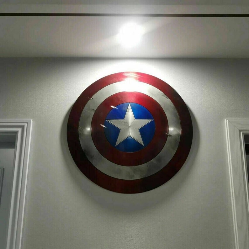 May include: A red, white, and blue Captain America shield with a white star on a blue circle in the center. The shield is hanging on a white wall.