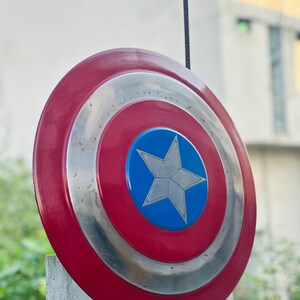 K&ouml;nnte beinhalten: Ein rot-wei&szlig;-blauer Captain America Schild mit einem silbernen Stern in der Mitte. Der Schild ist aus Metall und hat einen abgenutzten Look.