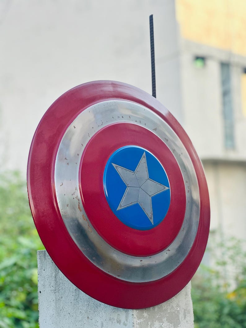 K&ouml;nnte beinhalten: Ein rot-wei&szlig;-blauer Captain America Schild mit einem silbernen Stern in der Mitte. Der Schild ist aus Metall und hat einen abgenutzten Look.