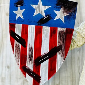 May include: A red, white, and blue shield with black paint splatters. The shield has three white stars on a blue background and red and white stripes on the bottom. The text "Iron Clad Armory" is written in black on the shield.