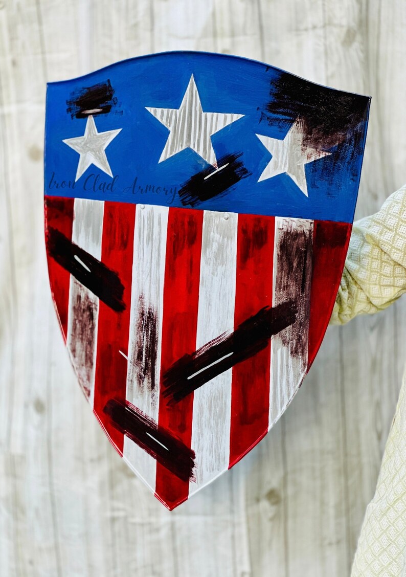 May include: A red, white, and blue shield with black paint splatters. The shield has three white stars on a blue background and red and white stripes on the bottom. The text "Iron Clad Armory" is written in black on the shield.