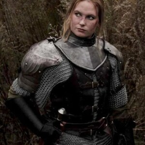 May include: A woman wearing medieval armor, including chainmail, a leather breastplate, and a metal helmet. She is standing in a forest setting.
