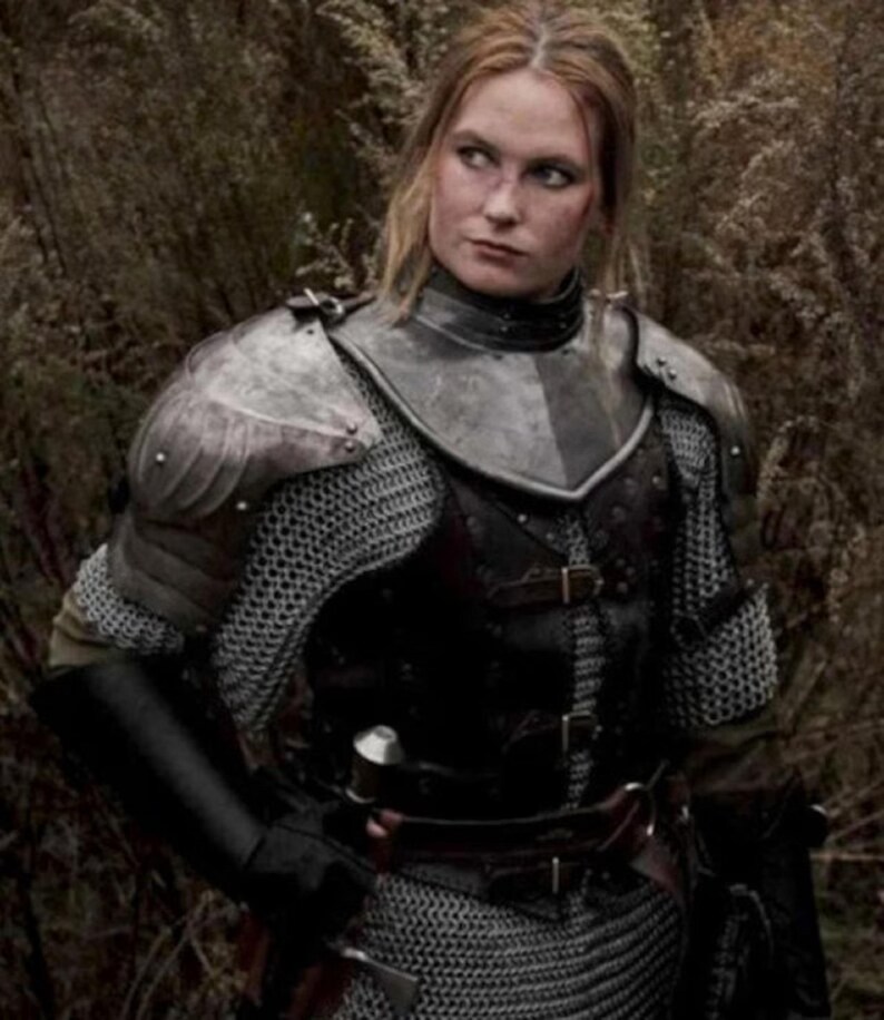 May include: A woman wearing medieval armor, including chainmail, a leather breastplate, and a metal helmet. She is standing in a forest setting.