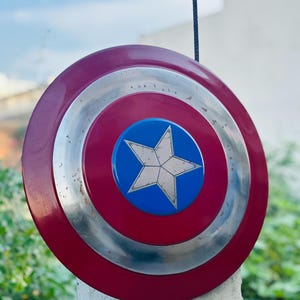K&ouml;nnte beinhalten: Ein rot-wei&szlig;-blauer Captain-America-Schild mit einem silbernen Metallrand. Der Schild ist auf einem wei&szlig;en Pfosten aufgestellt.