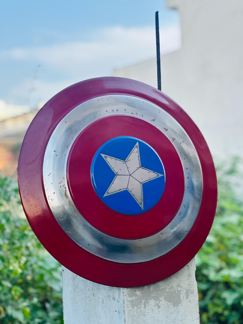K&ouml;nnte beinhalten: Ein rot-wei&szlig;-blauer Captain-America-Schild mit einem silbernen Metallrand. Der Schild ist auf einem wei&szlig;en Pfosten aufgestellt.
