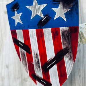 May include: A red, white, and blue shield with black paint splatters. The shield has a blue top with two white stars and a red, white, and blue striped bottom. The text "Iron Clad Armory" is written in white on the shield.