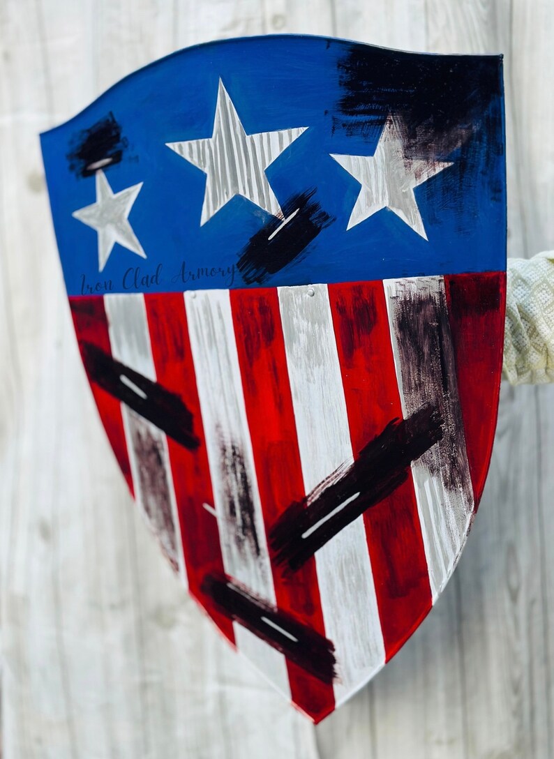 May include: A red, white, and blue shield with black paint splatters. The shield has a blue top with two white stars and a red, white, and blue striped bottom. The text "Iron Clad Armory" is written in white on the shield.