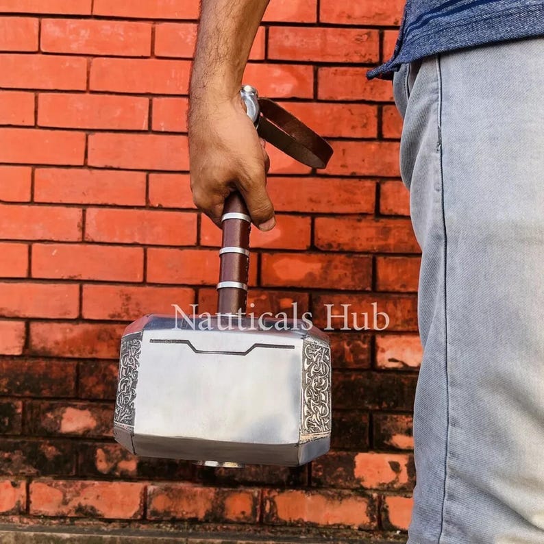 May include: A silver-colored hammer with a brown leather strap and intricate details. The hammer has a rectangular head with a raised center section and a textured surface. The hammer is held by a person's hand.