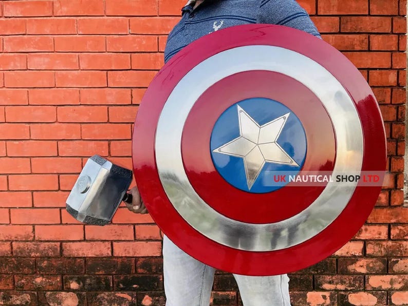May include: A person holding a large red, white, and blue Captain America shield with a silver star in the center. The shield has a silver rim and is made of metal. The person is also holding a silver and black hammer.