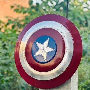 K&ouml;nnte beinhalten: Ein rot-wei&szlig;-blauer Captain America Schild mit einem f&uuml;nfzackigen Stern in der Mitte. Der Schild ist aus Metall und hat einen abgenutzten Look.