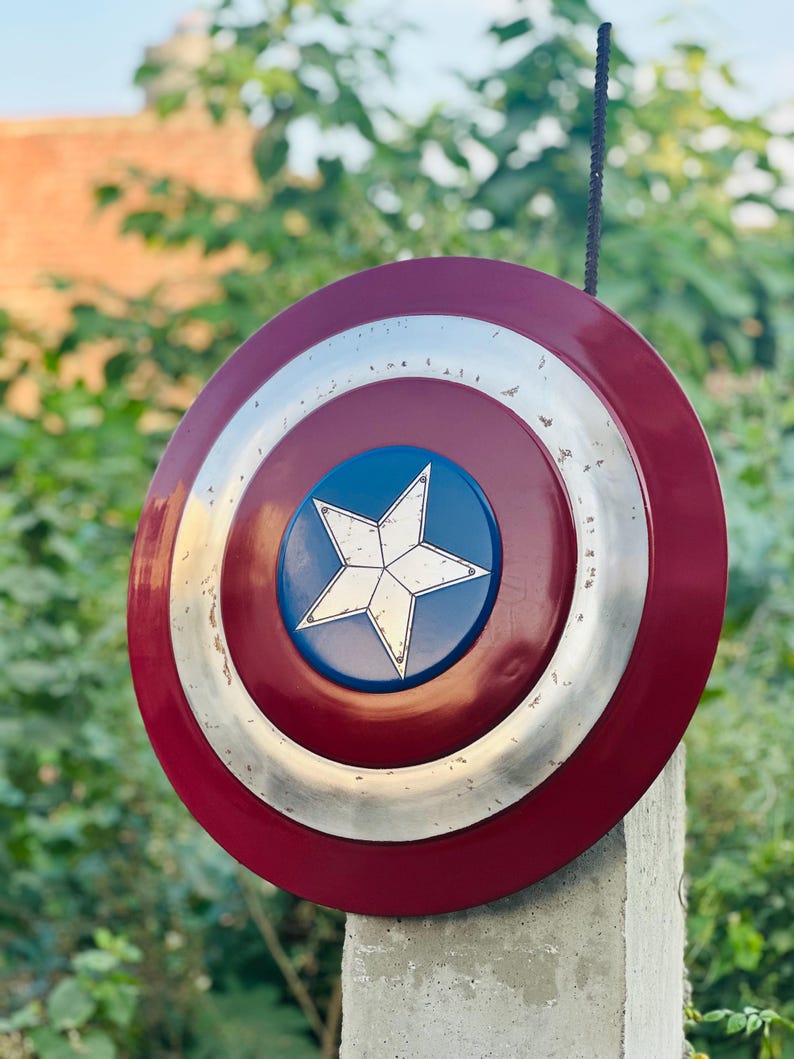 K&ouml;nnte beinhalten: Ein rot-wei&szlig;-blauer Captain America Schild mit einem f&uuml;nfzackigen Stern in der Mitte. Der Schild ist aus Metall und hat einen abgenutzten Look.