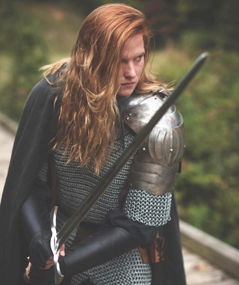 May include: A woman in chainmail armor and a black cloak holds a silver sword in front of her. She has long, red hair and is looking intently at something off-camera.