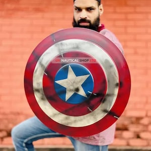 May include: A red, white, and blue Captain America shield with a distressed, weathered look. The shield has a five-pointed star in the center and is held by a person. The text "UK NAUTICAL SHOP LTD" is visible on the shield.