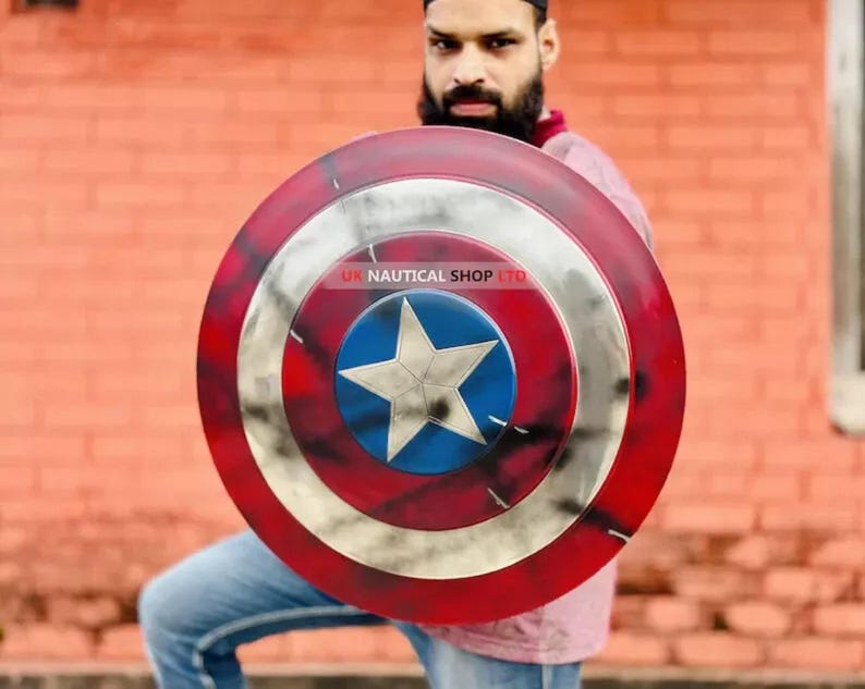 May include: A red, white, and blue Captain America shield with a distressed, weathered look. The shield has a five-pointed star in the center and is held by a person. The text "UK NAUTICAL SHOP LTD" is visible on the shield.