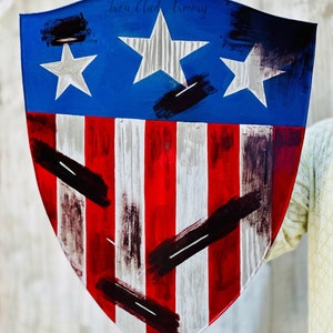 May include: A red, white, and blue shield with a distressed paint finish. The shield has three white stars on a blue background and red and white stripes. The shield is held by a hand and has the text "Iron Clad Armory" on the top.