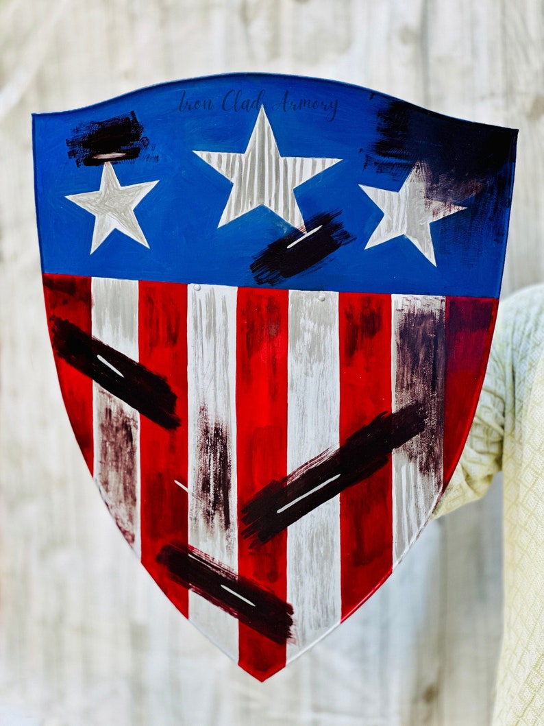 May include: A red, white, and blue shield with a distressed paint finish. The shield has three white stars on a blue background and red and white stripes. The shield is held by a hand and has the text "Iron Clad Armory" on the top.