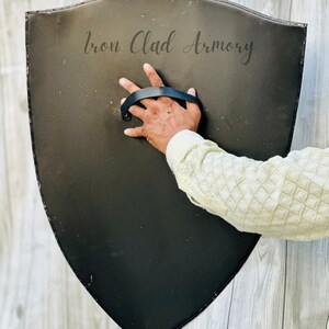 May include: A black, shield-shaped object with a leather handle. The shield is made of metal and has the words "Iron Clad Armory" printed on it.